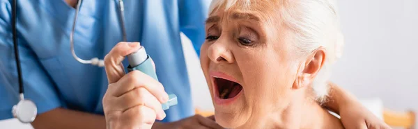 Donna anziana con bocca aperta affetta da attacco d'asma e con inalatore vicino all'infermiera su sfondo sfocato, striscione — Foto stock