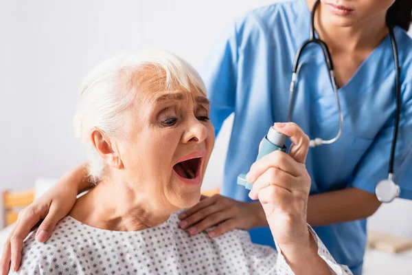 Femme âgée utilisant un inhalateur tout en souffrant d'une crise d'asthme près de l'infirmière touchant ses épaules sur fond flou — Photo de stock