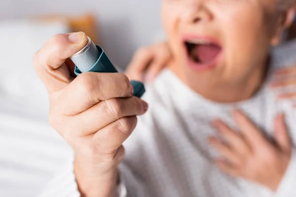 Vista ritagliata della donna anziana con la bocca aperta che tiene l'inalatore mentre soffre di attacco di asma, sfondo sfocato — Foto stock