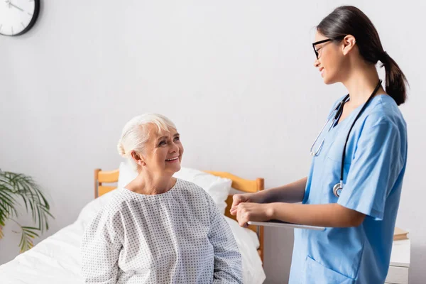 Giovane infermiera asiatica con tablet digitale mentre parla con sorridente donna anziana in ospedale — Foto stock