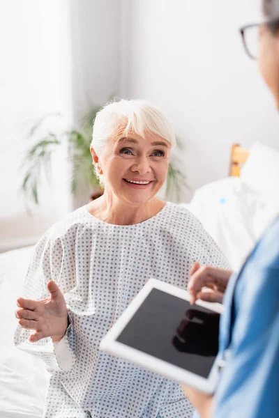 Mujer anciana feliz gesto mientras habla con la enfermera sosteniendo tableta digital con pantalla en blanco en primer plano borrosa - foto de stock