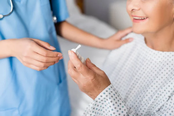 Cropped view of aged, smiling woman holding nasal spray near nurse on blurred background — Stock Photo