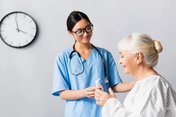 Smiling asian nurse giving nasal spray to senior woman in hospital — Stock Photo