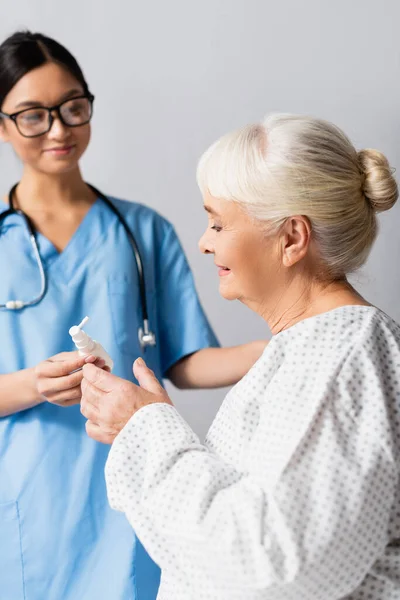 Молода азіатська медсестра дає носовий спрей старшій жінці, розмитий фон — стокове фото