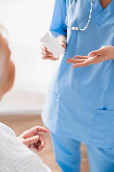 Vue partielle de l'infirmière pointant le récipient avec des médicaments près de la femme âgée au premier plan flou — Photo de stock