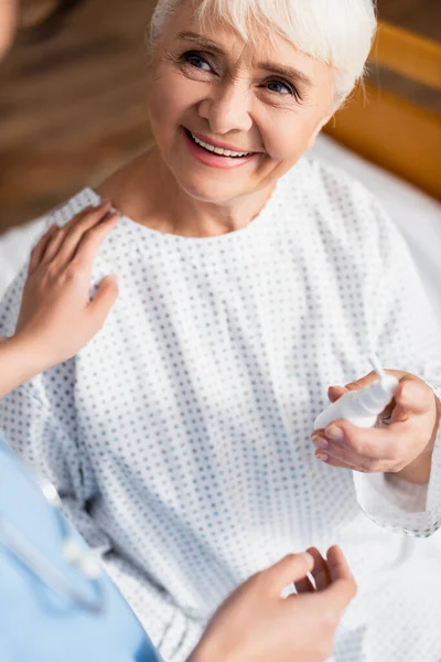 Sonriente mujer mayor sosteniendo aerosol nasal cerca de enfermera tocando escuchar hombro en primer plano borrosa - foto de stock