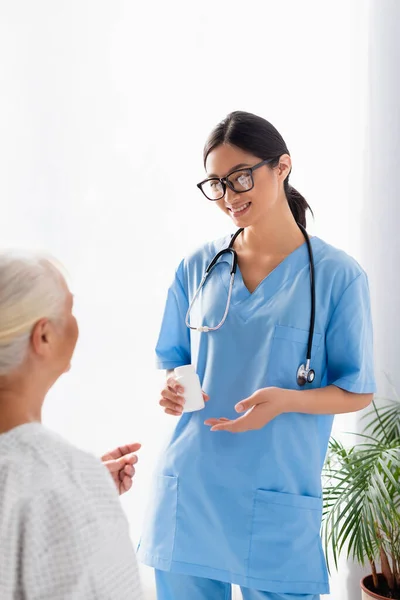 Smiling asian woman holding container with pills near senior woman on blurred foreground — Stock Photo