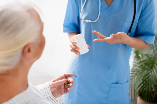 Teilaufnahme einer Krankenschwester, die auf einen Behälter mit Medikamenten in der Nähe einer betagten Frau zeigt, im unscharfen Vordergrund — Stockfoto