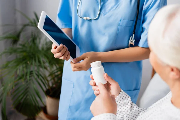 Vista ritagliata di donna anziana che punta con il dito al contenitore pillole vicino infermiera in possesso di tablet digitale — Foto stock