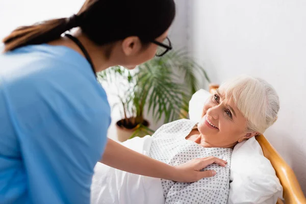 Junge Krankenschwester berührt fröhliche alte Frau, die im Krankenhaus liegt, verschwommener Vordergrund — Stockfoto