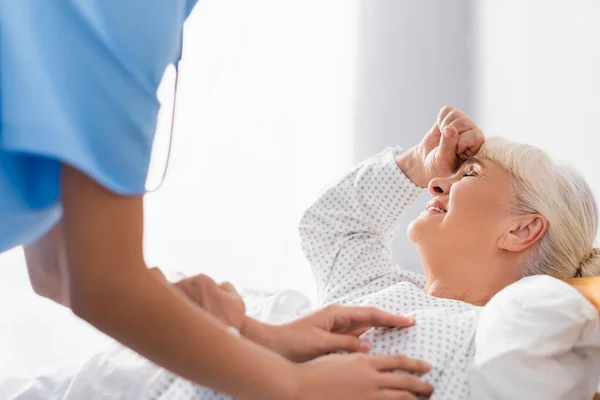 Infirmière toucher une femme âgée souffrant de maux de tête tout en étant couché les yeux fermés, avant-plan flou — Photo de stock