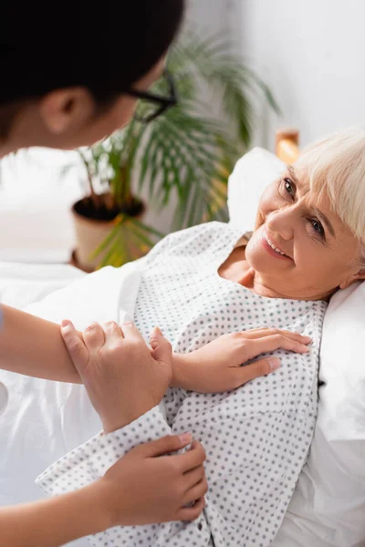 Femme âgée souriante regardant infirmière la toucher sur le premier plan flou — Photo de stock