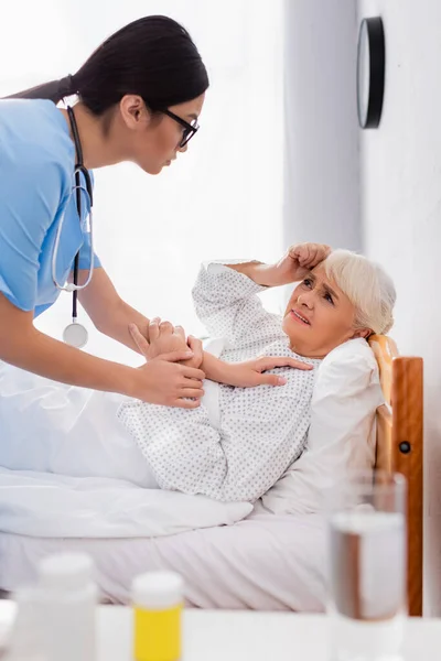Joven asiático mujer tocando enfermo senior mujer acostado en cama y sufriendo de dolor de cabeza, borrosa primer plano - foto de stock