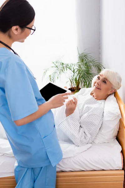 Mulher smiling sênior falando com jovem enfermeira segurando tablet digital em primeiro plano turvo — Fotografia de Stock