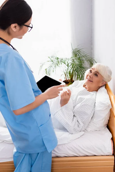 Lächelnde Seniorin im Gespräch mit junger Krankenschwester mit digitalem Tablet im verschwommenen Vordergrund — Stockfoto