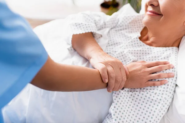 Vista recortada de la enfermera tocando sonriente mujer mayor acostada en el hospital, borrosa primer plano - foto de stock