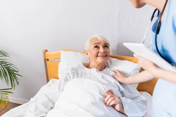 Junge Krankenschwester mit digitalem Tablet und berührender Schulter einer älteren Frau, die im Bett liegt, verschwommener Vordergrund — Stockfoto