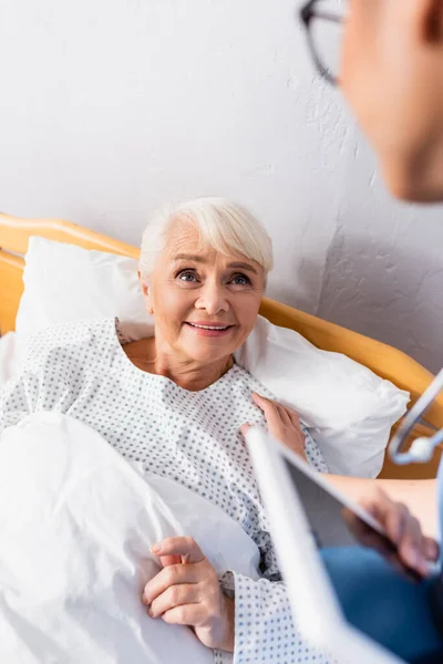 Nurse with digital tablet touching shoulder of smiling aged woman lying in bed, blurred foreground — Stock Photo