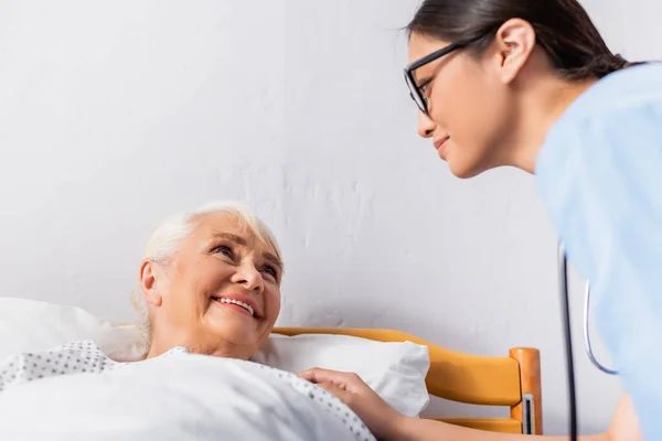 Young asian nurse looking at smiling elderly woman lying in hospital — Stock Photo