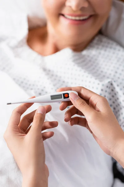 Partial view of nurse holding thermometer near smiling senior woman on blurred background — Stock Photo