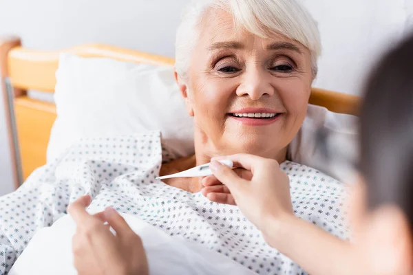 Обрезанный вид улыбающейся пожилой женщины рядом с медсестрой с термометром на размытом переднем плане — стоковое фото