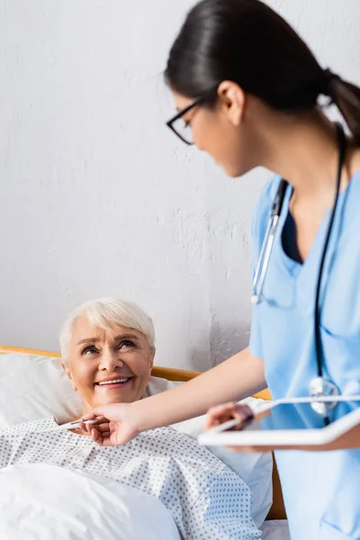 Junge asiatische Krankenschwester gibt Thermometer an fröhliche Seniorin, verschwommener Vordergrund — Stockfoto