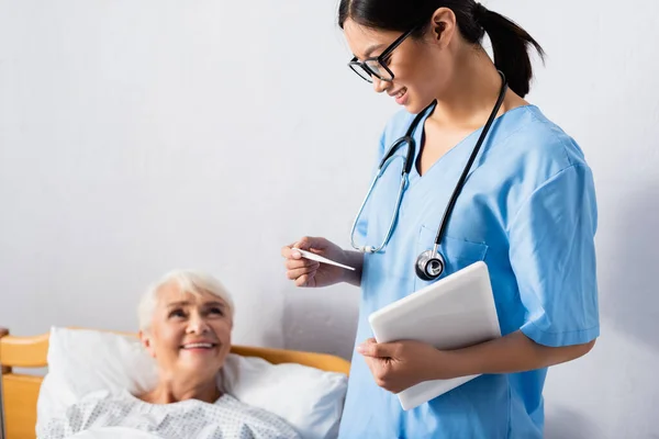 Smiling asian nurse with digital tablet looking at thermometer near happy elderly woman on blurred background — Stock Photo