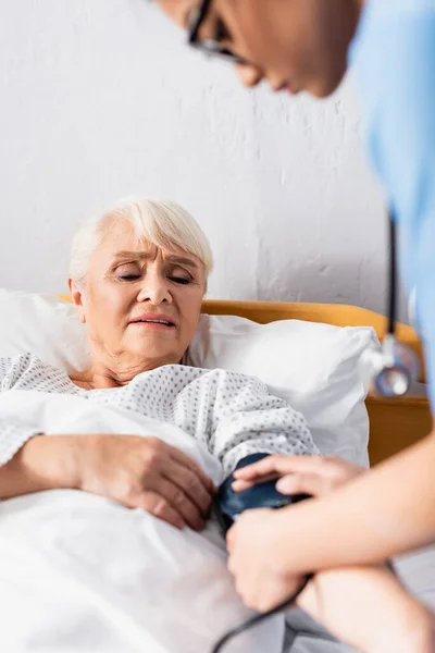 Asian nurse fixing cuff of tonometer on arm of aged woman, blurred foreground — Stock Photo