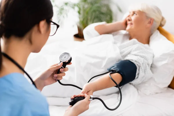 Mise au point sélective du tonomètre dans les mains d'une jeune infirmière mesurant la pression artérielle d'une femme âgée souffrant de maux de tête — Photo de stock