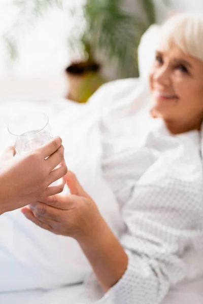 Infirmière donnant un verre d'eau à une femme âgée souriante à l'hôpital, fond flou — Photo de stock