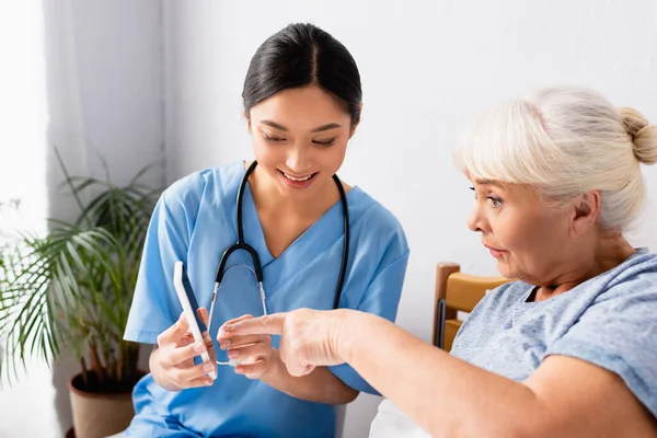 Astonished elderly woman pointing with finger at mobile phone in hands of smiling asian nurse — Stock Photo