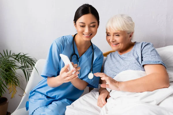 Веселая азиатская медсестра с помощью мобильного телефона рядом счастливая пожилая женщина лежит в постели — стоковое фото