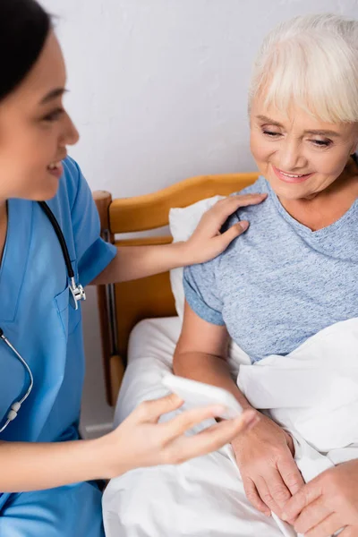 Молодая азиатская медсестра показывает смартфон улыбающейся пожилой женщине в больнице, размытый передний план — стоковое фото
