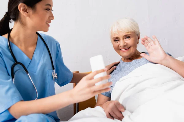 Молодая азиатская медсестра держит мобильный телефон рядом с веселой пожилой женщиной, машущей рукой во время видеозвонка — стоковое фото