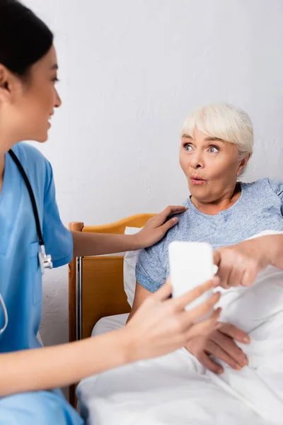 Espantado idosa mulher apontando para smartphone perto jovem asiático enfermeira, foreground borrado — Fotografia de Stock