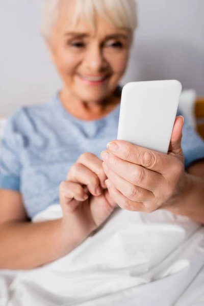 Cheerful elderly woman chatting on smartphone in hospital, blurred background — Stock Photo