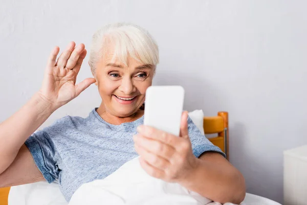 Heureuse femme âgée agitant la main pendant le chat vidéo sur smartphone à l'hôpital — Photo de stock