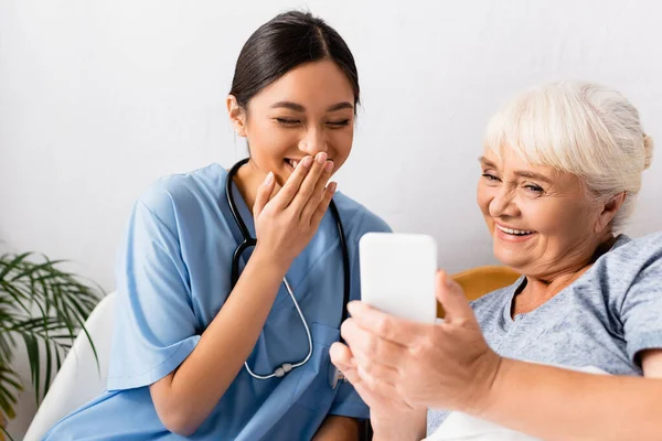 Смеющаяся азиатская медсестра, прикрывающая рот рукой рядом с веселой пожилой женщиной с помощью мобильного телефона, размытый передний план — стоковое фото