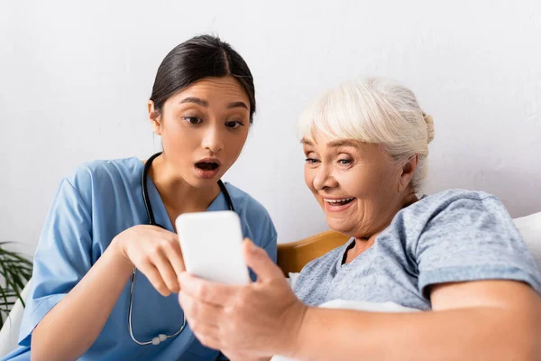 Изумленная азиатская медсестра, указывающая на мобильный телефон рядом с взволнованной пожилой женщиной, размытый передний план — стоковое фото