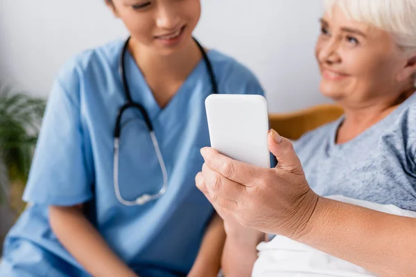 Cheerful senior woman showing mobile phone to smiling asian nurse on blurred background — Stock Photo