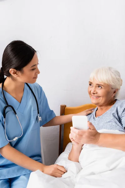 Счастливая пожилая женщина смотрит на улыбающуюся азиатскую медсестру, держа смартфон с чистым экраном — стоковое фото