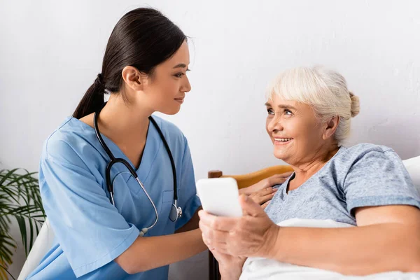 Усміхнена азіатська медсестра торкається плеча щасливої старшої жінки, використовуючи смартфон у лікарні — стокове фото