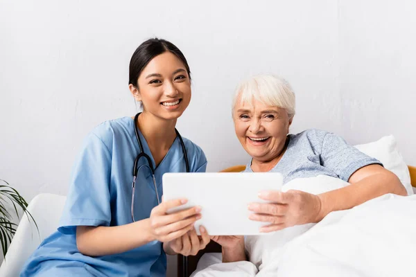 Happy asian nurse and laughing senior woman looking at camera while holding digital tablet — Stock Photo