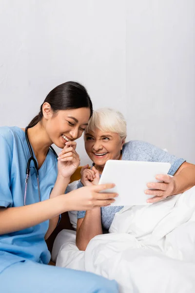 Aufgeregte asiatische Krankenschwester und Seniorin lachen, während sie gemeinsam ein digitales Tablet benutzen — Stockfoto