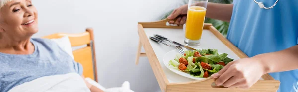 Heureux Eldrely femme regardant infirmière tenant plateau avec petit déjeuner, fond flou, bannière — Photo de stock