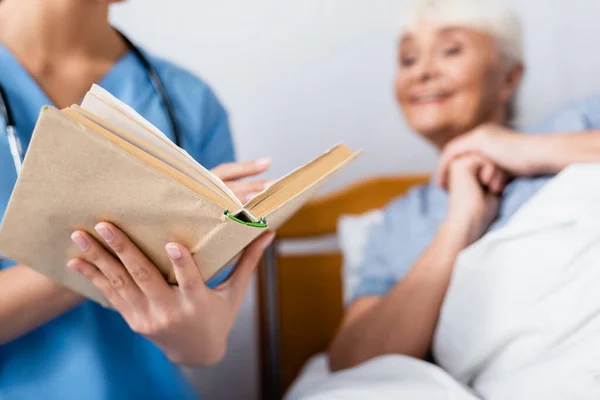 Infirmière lecture livre à excité femme âgée à la clinique, flou au premier plan — Photo de stock