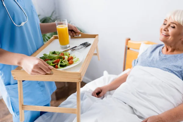 Nurse holding tray with fresh breakfast near pleased senior woman, blurred background — Stock Photo