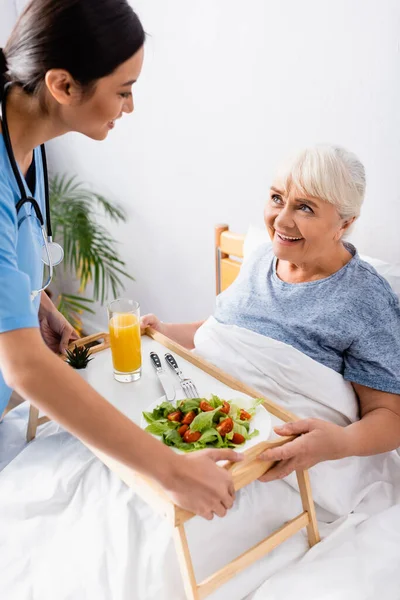 Sonriente asiático enfermera celebración bandeja con desayuno cerca feliz anciano mujer en cama - foto de stock