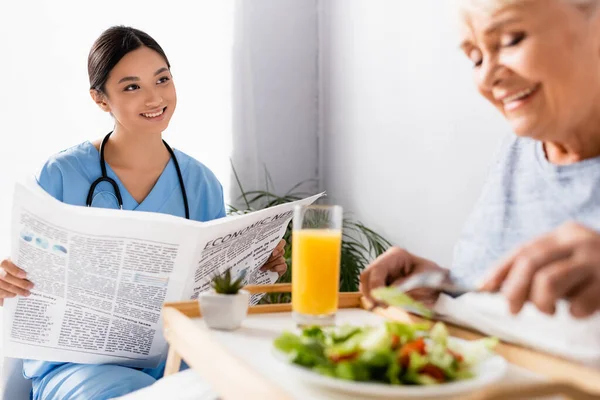 Sorrindo asiático enfermeira segurando jornal perto satisfeito sênior mulher tendo café da manhã no embaçado primeiro plano — Fotografia de Stock