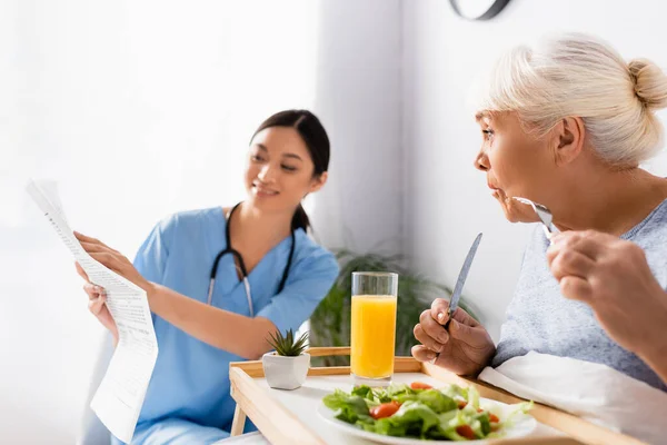 Amazed senior woman having breakfast and looking at smiling asian nurse reading newspaper on blurred background — Stock Photo